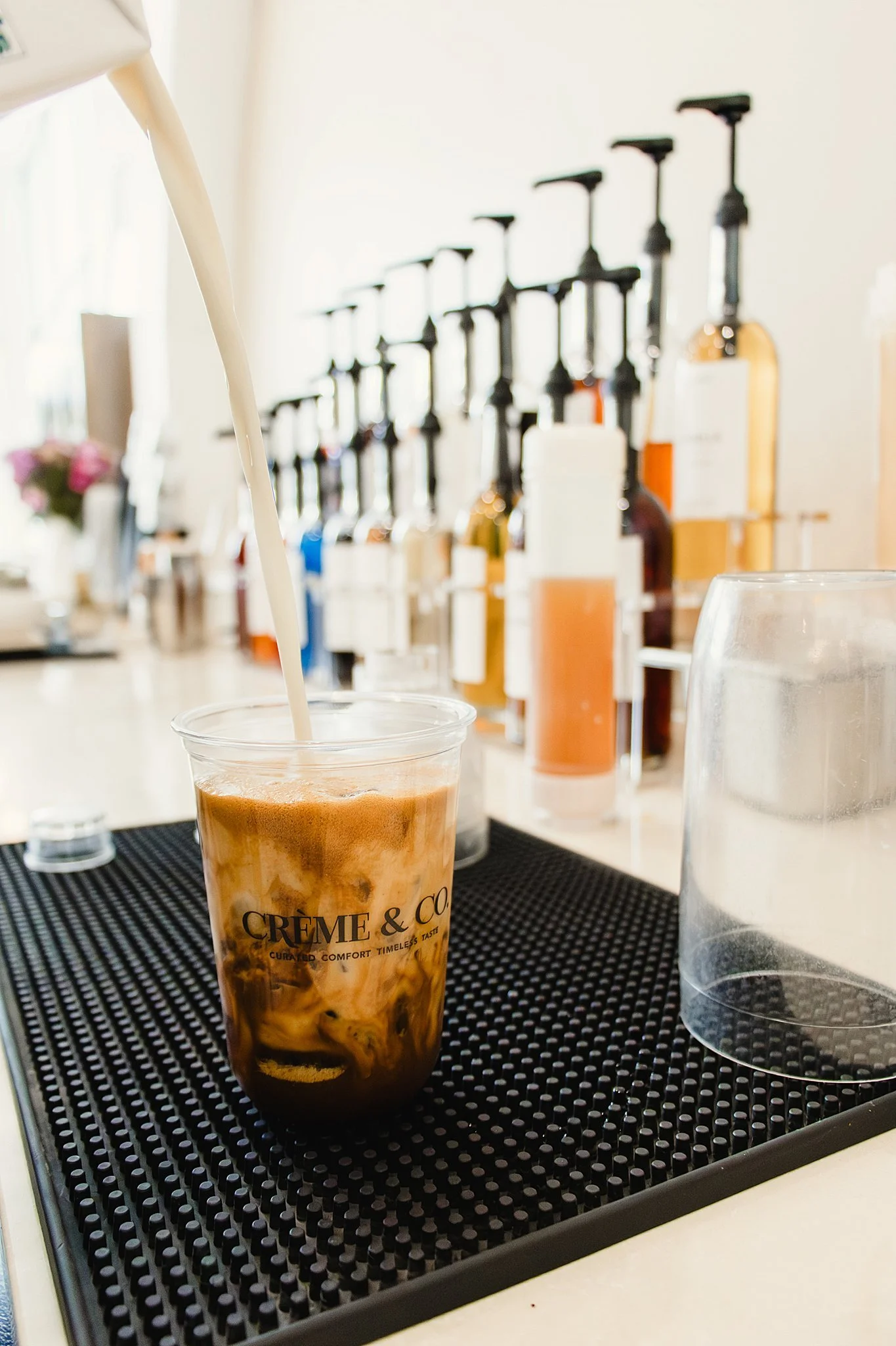 Close-up of a plastic cup filled with iced coffee with milk, placed on a black rubber bar mat, with a row of various liquor bottles with pump dispensers in the background with Naperville branding photographer.