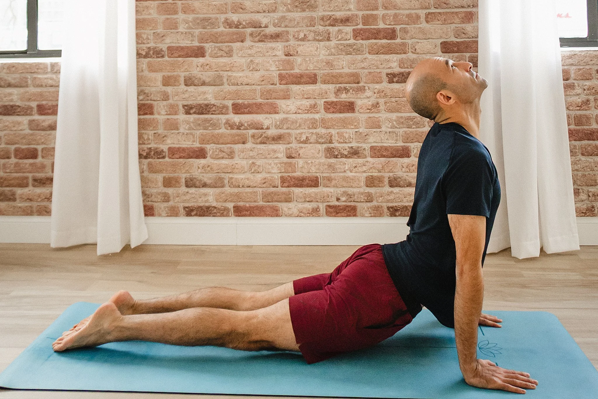 A man doing yoga on a blue mat in a room with brick walls and white curtains, in a cobra pose with his head tilted back and looking up for small business branding photos near me.