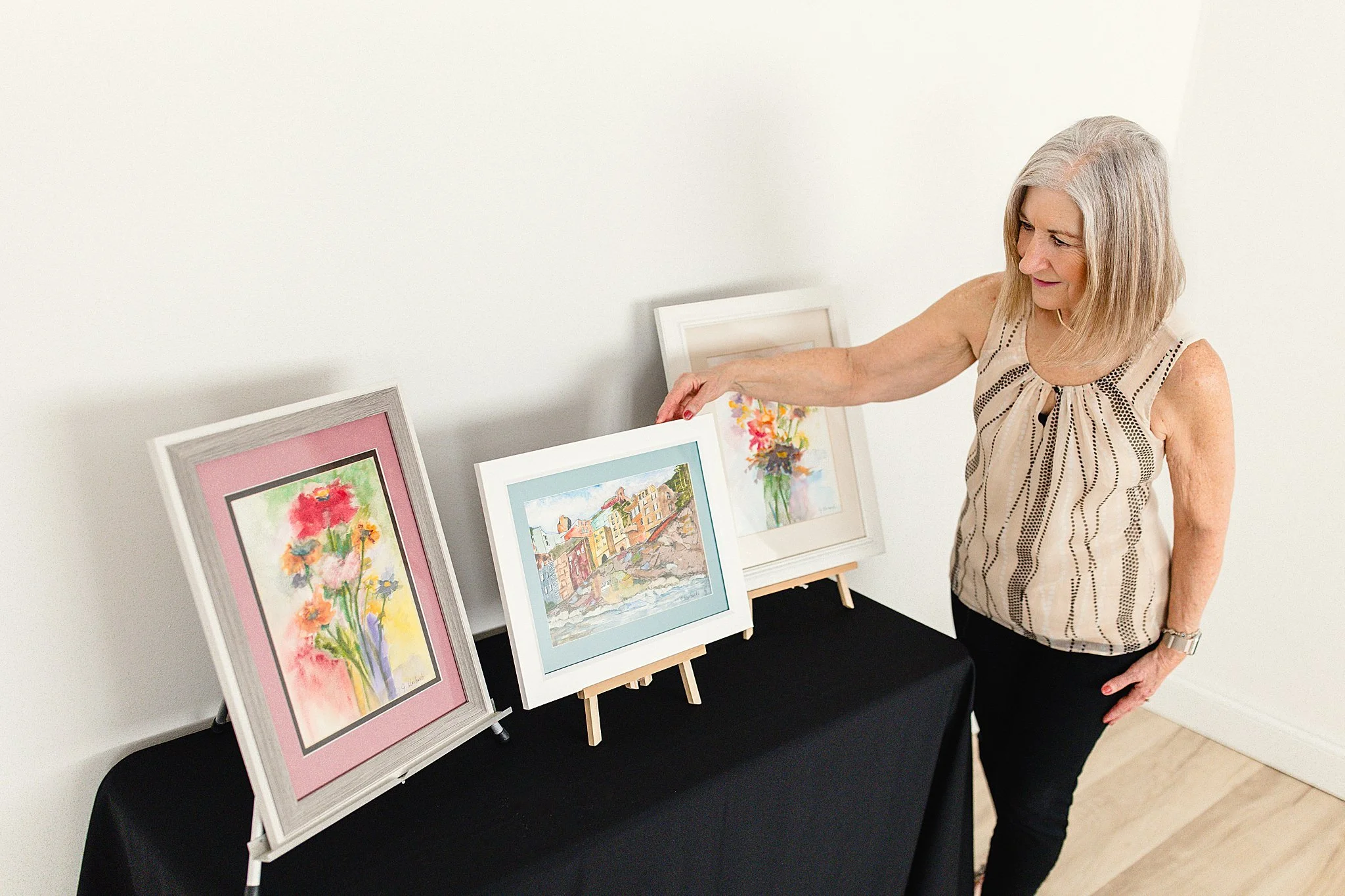 A woman with blonde hair looking at three framed watercolor paintings on display on a black table against a white wall with Naperville branding photographers.