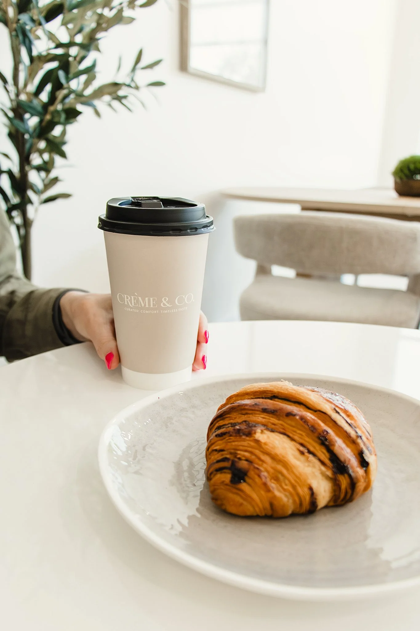 A hand holding a to-go coffee cup labeled 'CRÈME & CO.' with a croissant on a white plate in the foreground, inside a bright, modern cafe with light-colored walls and a large window during brand photography Naperville.