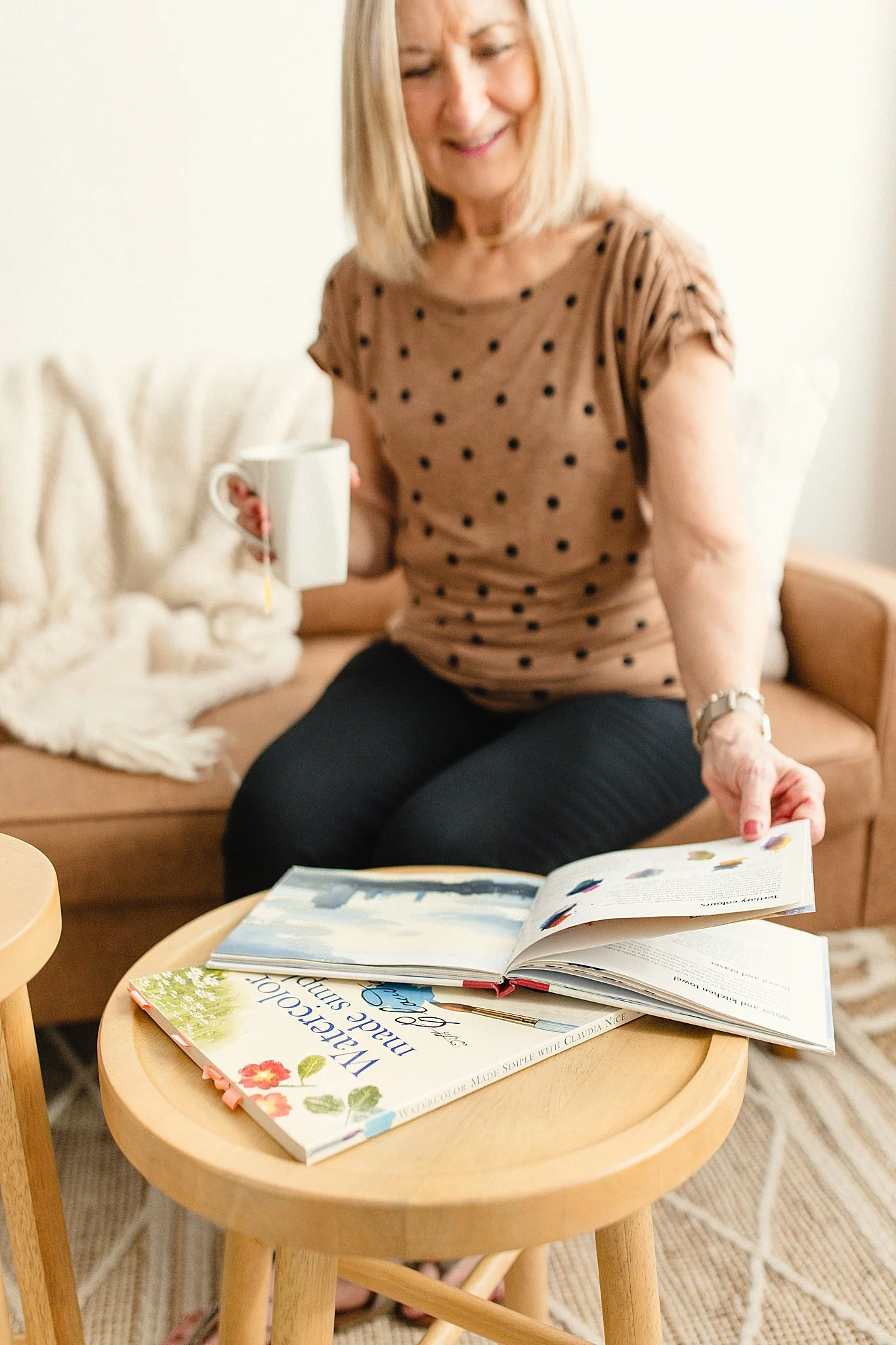 A woman sitting on a sofa, flipping through a colorful watercolor art book on a round wooden table while holding a white mug, with a fluffy white cat lying nearby for brand photographers in Naperville, IL.