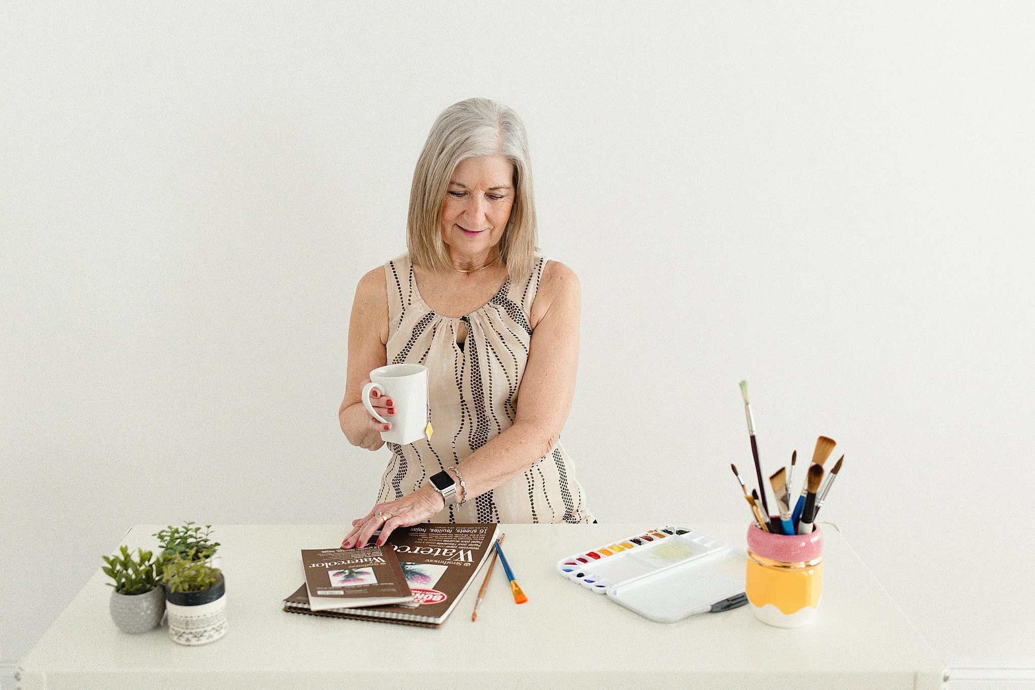 An elderly woman with gray hair, wearing a sleeveless patterned top, is standing at a white table. She is holding a white mug in one hand and resting her other hand on a magazine during a brand photoshoot in Naperville, IL with Ally and B Photography