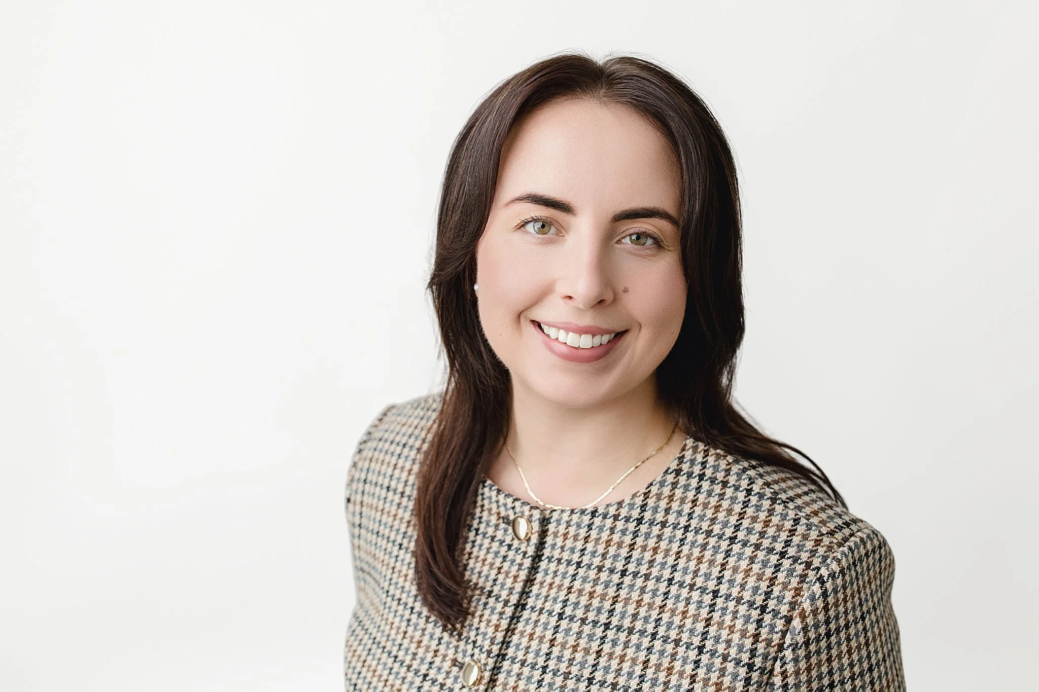 A woman with shoulder-length dark brown hair, wearing a checkered blazer with pearl buttons, smiling against a plain white background during a professional photography session with Naperville headshot photographer.