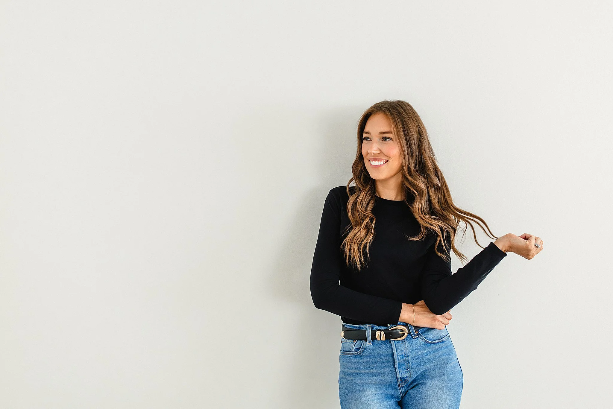 A young woman with long wavy brown hair, wearing a black long-sleeve shirt and blue jeans, standing against a plain white wall, smiling and looking to the side during a brand photo session in Naperville, IL.