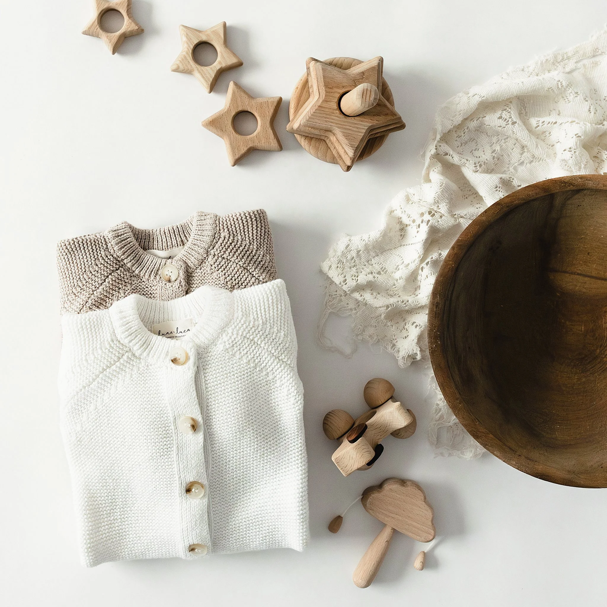 Two folded knitted sweaters, one beige and one white, placed on a white surface. Wooden toys including a star, a mushroom, and a car are scattered around. A large wooden bowl and a lace fabric are also visible for flat lays in Naperville.