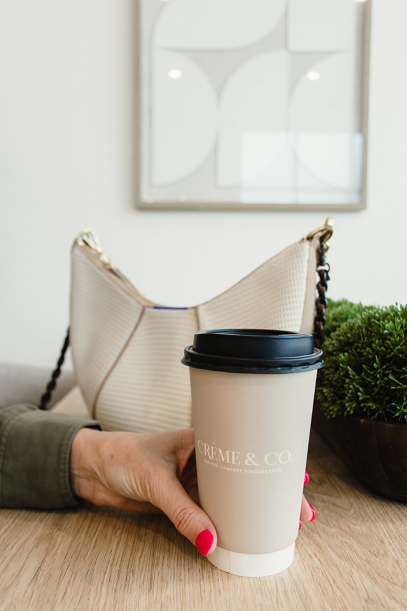 A person's hand with pink nail polish holding a white to-go coffee cup with a black lid labeled 'Crème & Co.' on a wooden table. In the background, a beige handbag, a green plant, and a wall mirror are visible for branding photography.