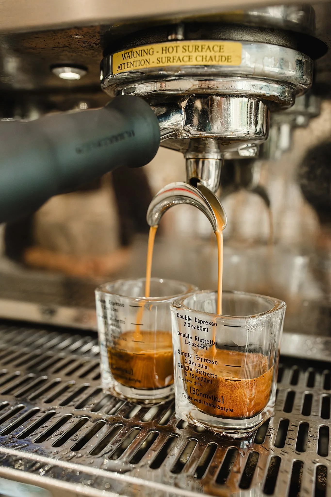 Close-up of espresso being poured from a coffee machine into glass measuring cups, with a warning sticker about hot surfaces on the machine for coffee shop branding photographers near me.