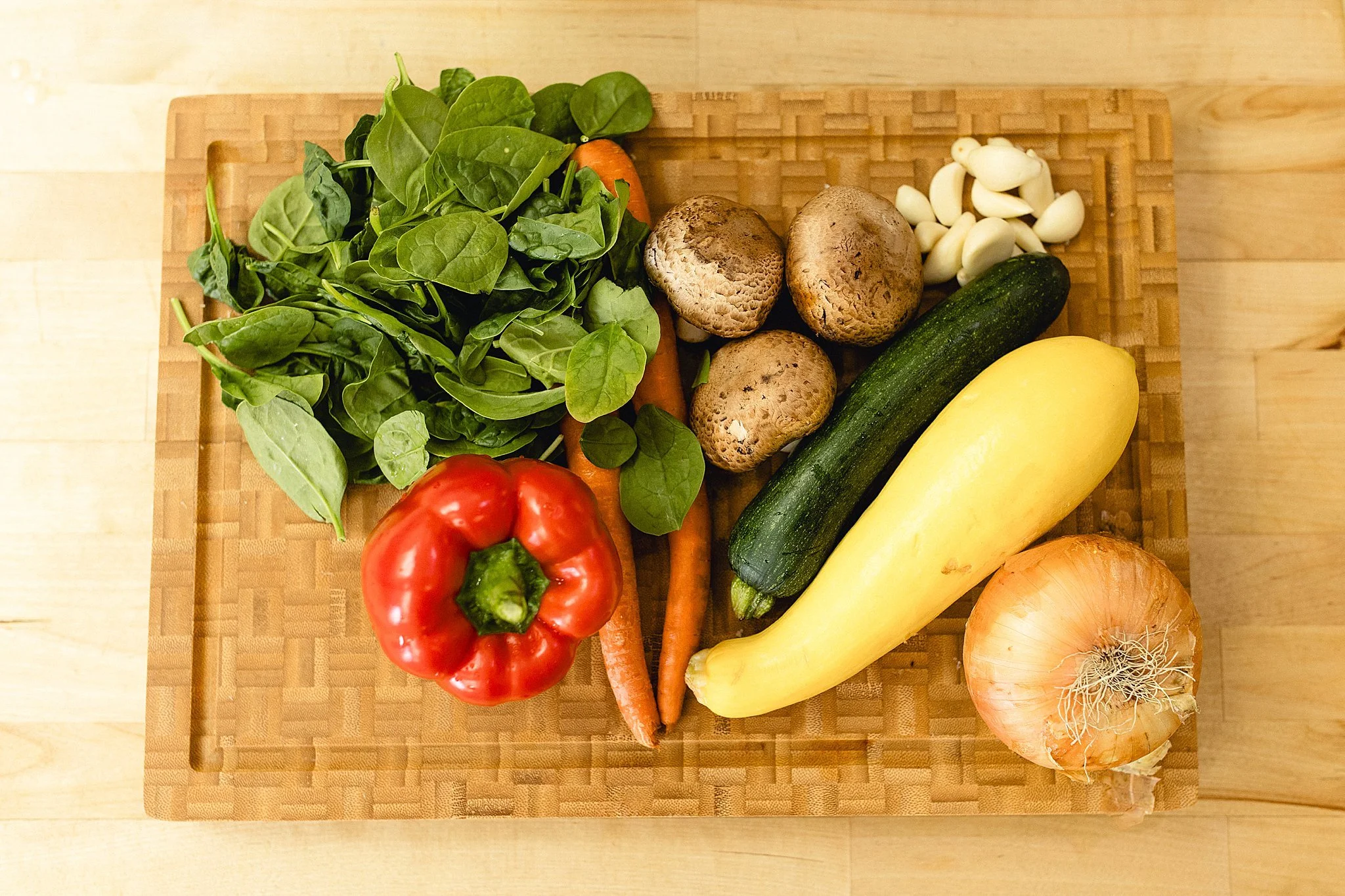 Fresh vegetables on a wooden cutting board, including spinach, a red bell pepper, carrots, mushrooms, garlic cloves, zucchini, yellow squash, and an onion for branding photos in Naperville, IL.