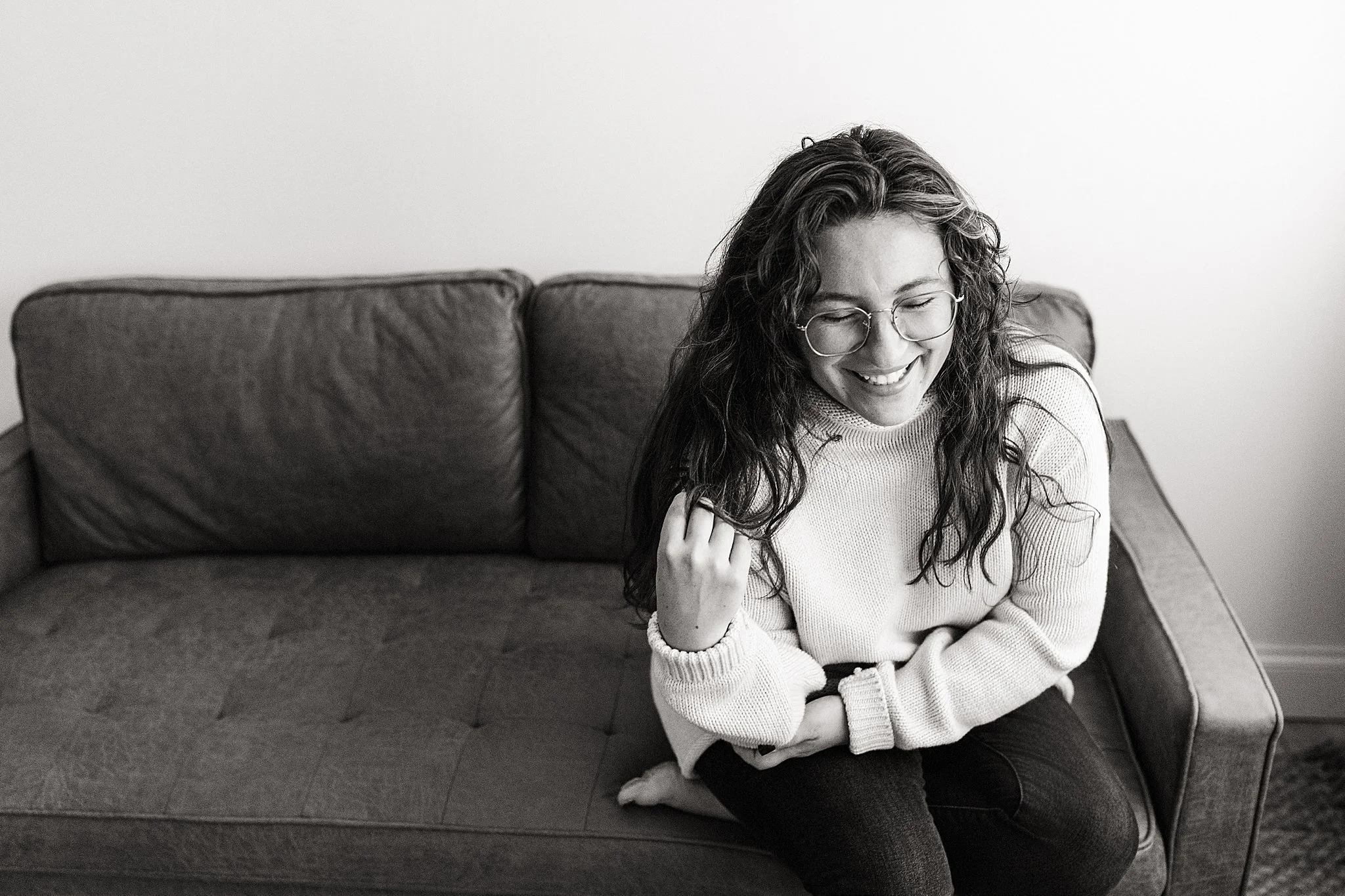 A woman with curly hair and glasses sitting on a couch, smiling and looking down with a Naperville brand photographer.