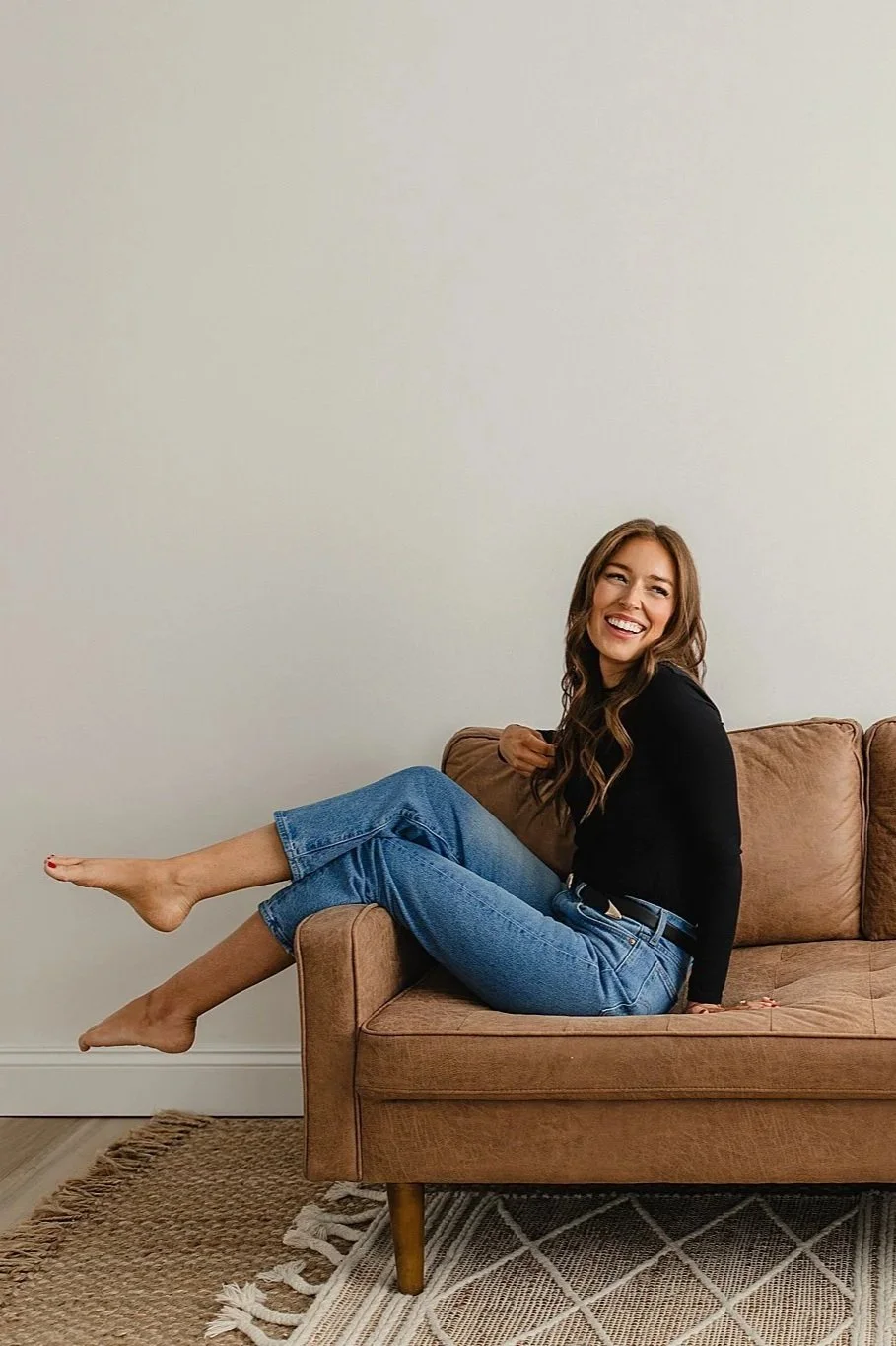A woman sitting on a brown couch, smiling, with her legs stretched out and toes touching the floor. She is wearing a black long-sleeve top and blue jeans, with long wavy brown hair for personal branding photography.