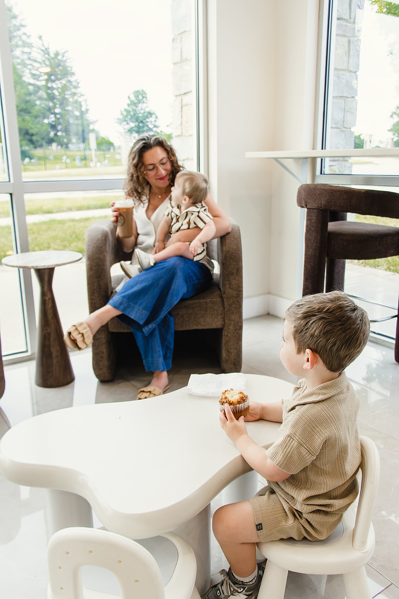 A woman with curly hair and glasses sitting in a lounge chair, holding a drink and looking at a young boy sitting nearby with a muffin in his hand. The woman is wearing beige sandals, a white top, and blue trousers. The boy is wearing a brown shirt a
