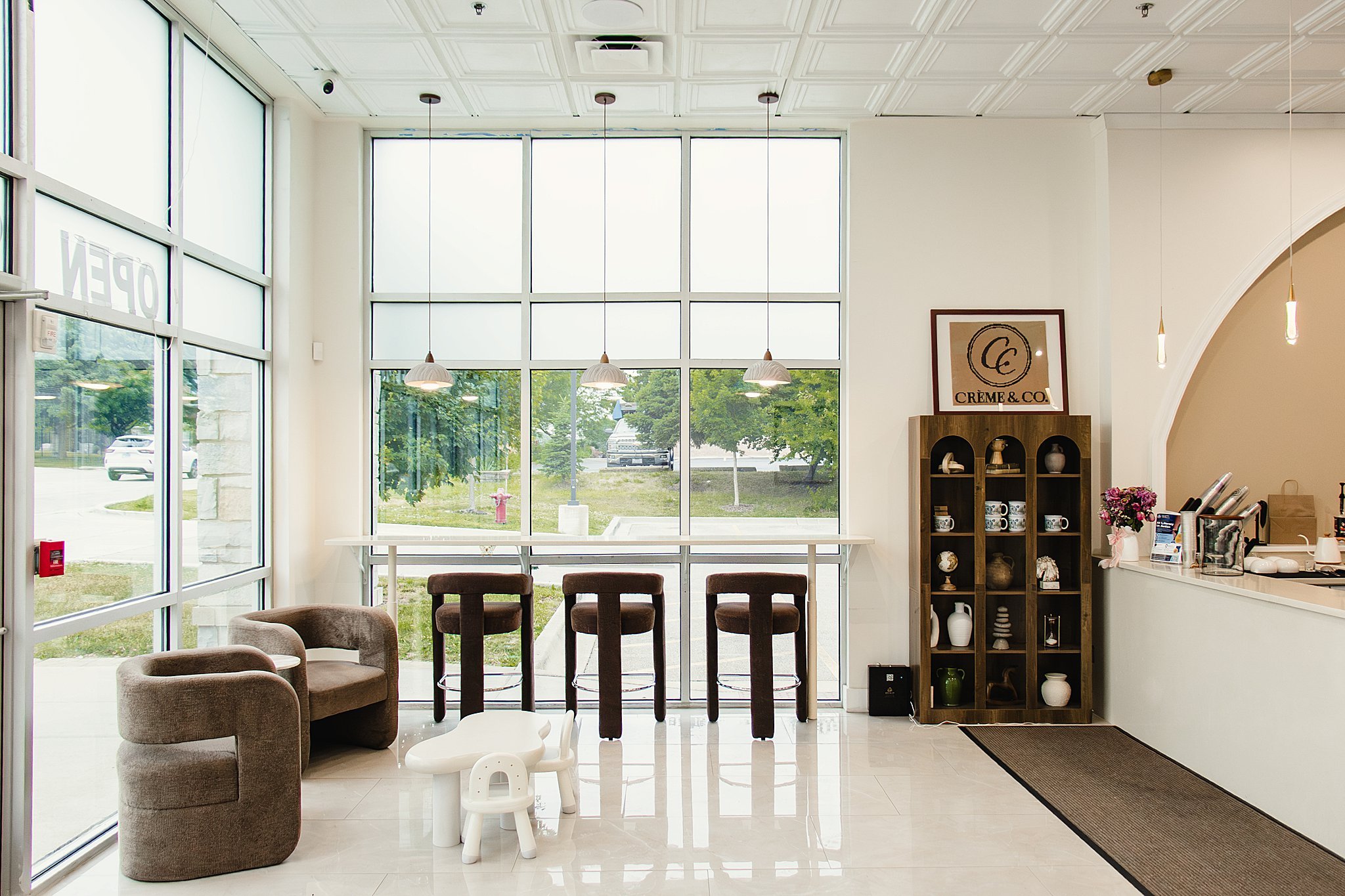 Interior of a modern cafe or waiting area with large floor-to-ceiling windows, a white tiled floor, and a variety of seating including armchairs and chairs. There are pendant lights hanging from the ceiling, a wooden display shelf with decorative ite