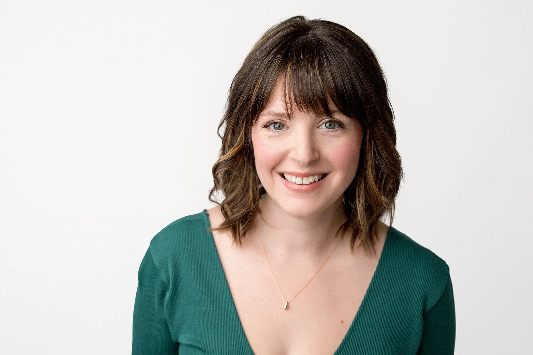 A young woman with shoulder-length brown hair, blue eyes, and a friendly smile, wearing a green V-neck top and a delicate gold necklace, against a plain white background during a Naperville headshot photoshoot near me.
