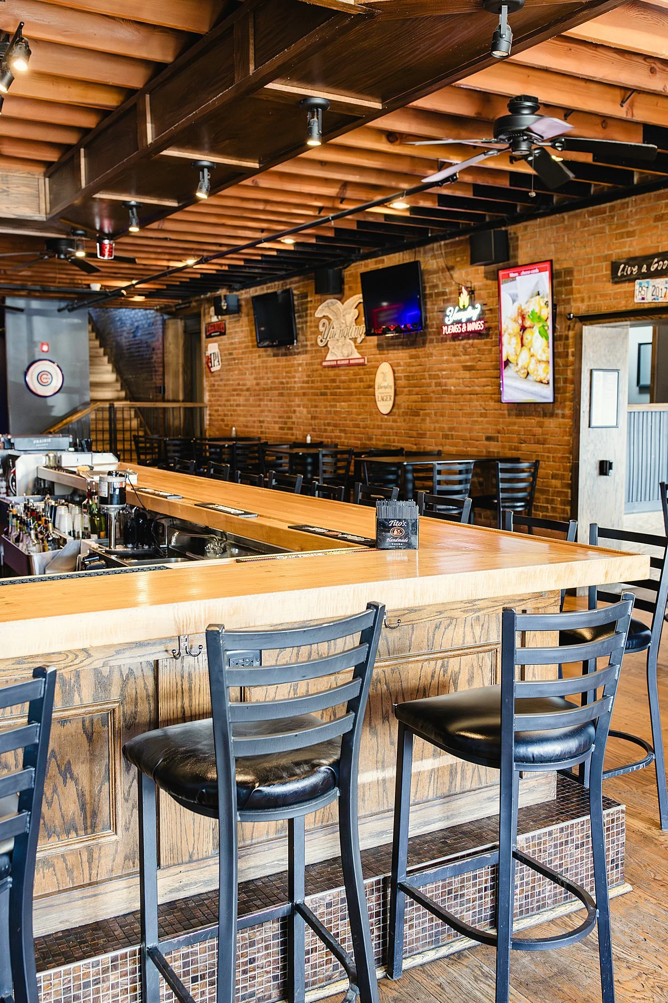 Interior of a bar with a wooden counter, black chairs, exposed brick walls, TV screens, and ceiling fans for brand photographers near me.