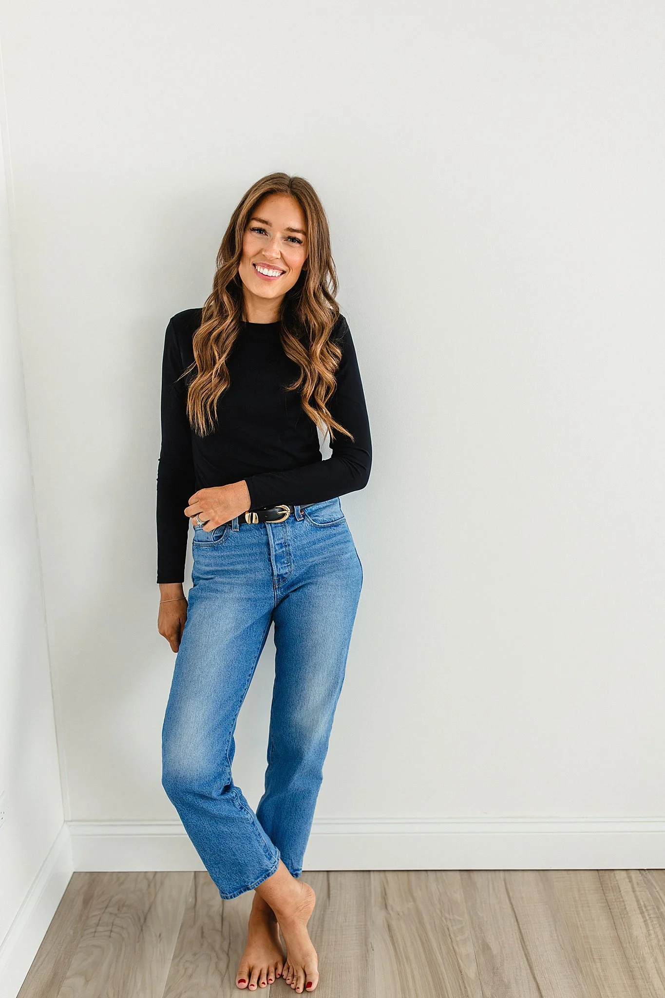 A woman with long wavy brown hair, wearing a black long-sleeve shirt and blue jeans, smiling and standing barefoot against a light-colored wall during Naperville brand photos.