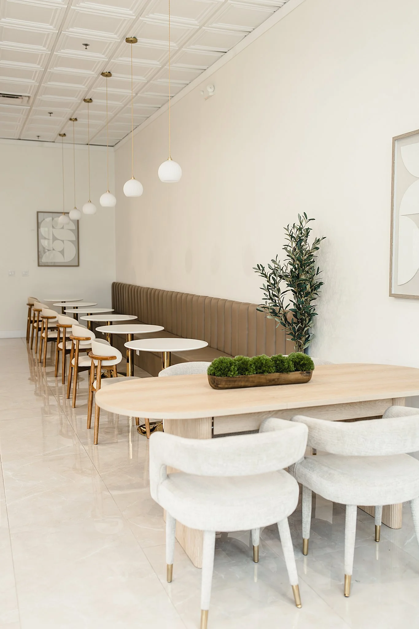 A modern and minimalistic dining area with a row of white tables and chairs, beige booth seating, a large potted plant, and decorative wall art, illuminated by pendant lights hanging from a white ceiling with branding photographer Naperville.