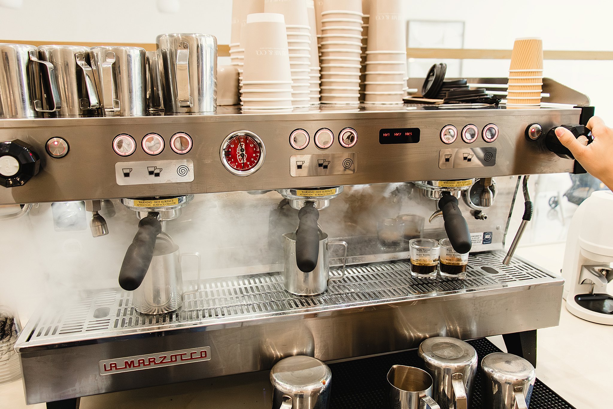 A La Marzocco espresso machine with stacks of paper cups and cups of espresso on a tray for a brand photoshoot near me.