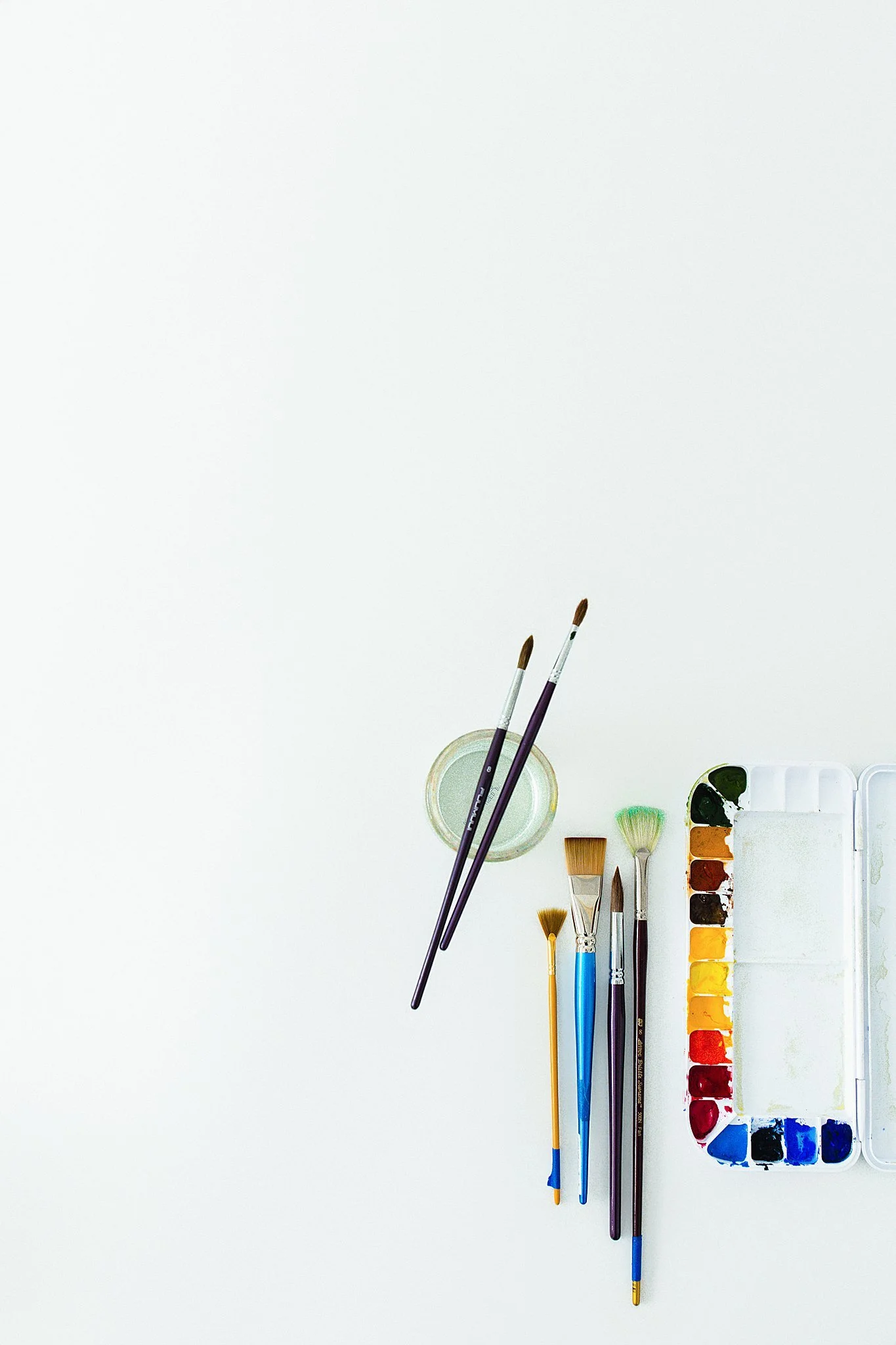 Paintbrushes, watercolor palette, and water container on a white surface for flat lay photographers near me.