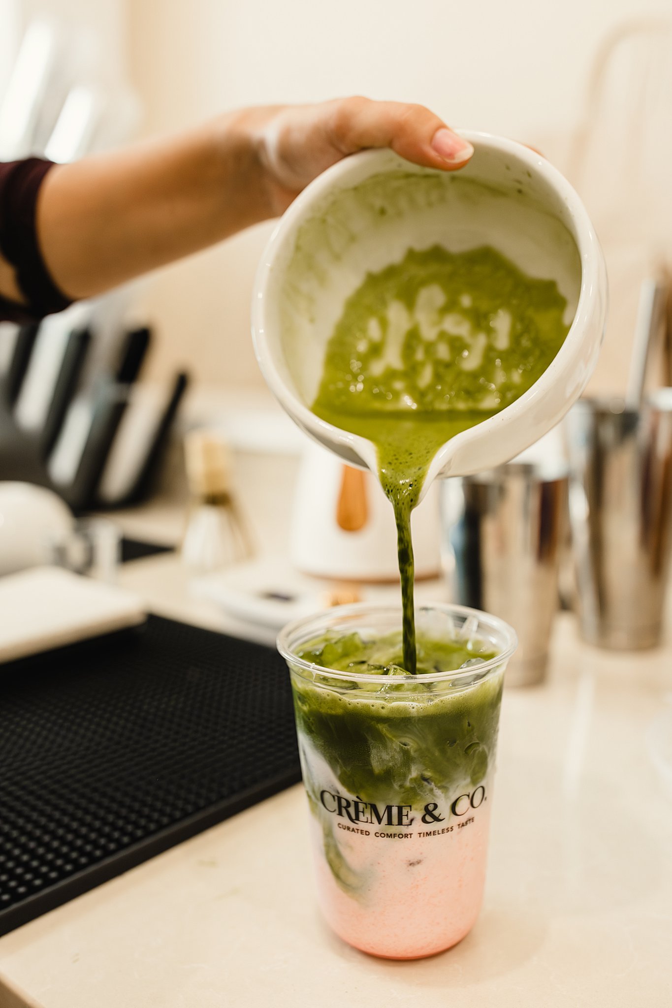 Green matcha latte being poured into a clear cup with pink-colored foam at the bottom, on a white countertop with kitchen utensils in the background with branding photographers in Naperville, IL.