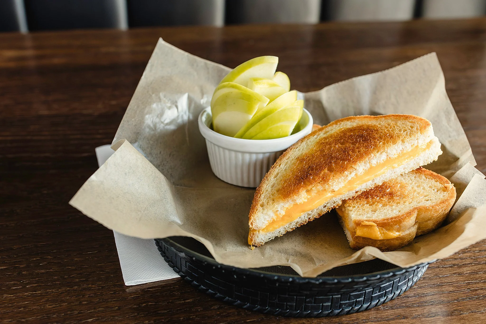 A grilled cheese sandwich cut in half with melted cheese, served with sliced green apples in a small white ramekin on a black basket lined with brown parchment paper on a wooden table with commercial food photographers near Naperville, IL.