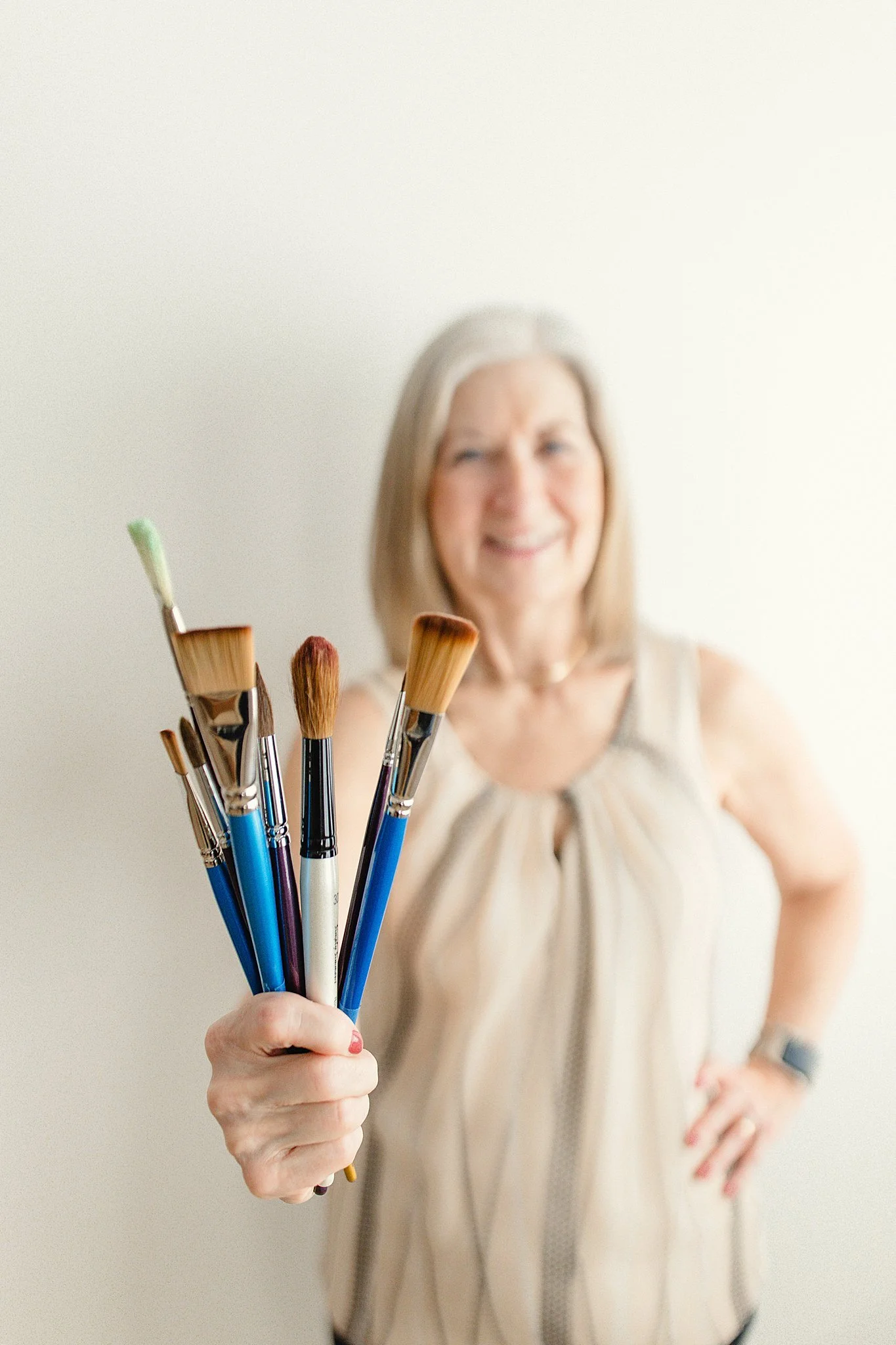 An older woman holding several paintbrushes towards the camera, smiling and standing against a plain white background for artist branding photos near me.