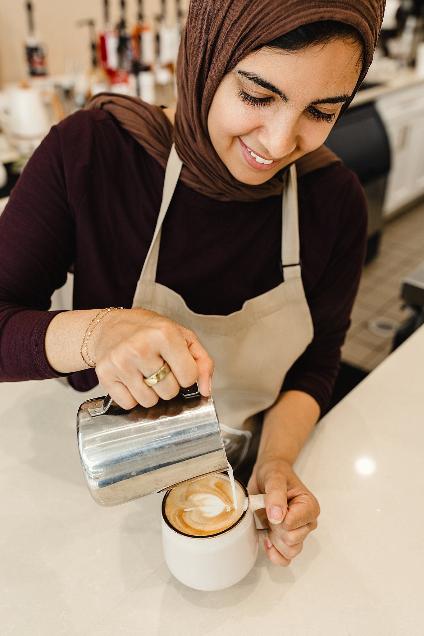 A woman wearing a hijab and apron is pouring steamed milk into a cup of coffee, creating latte art in a cafe setting for branding photographer in Naperville, IL.