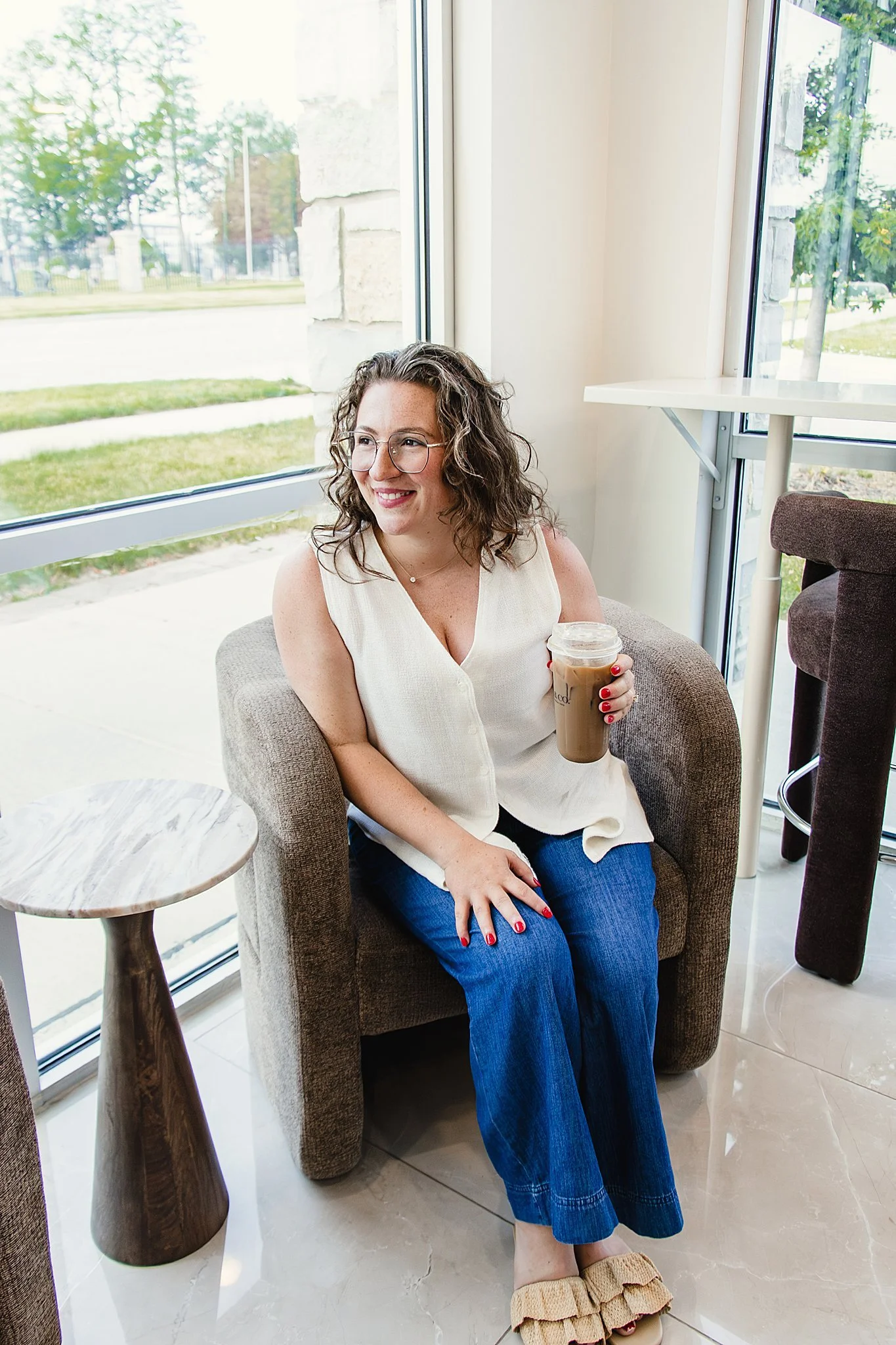 A woman with curly hair and glasses sitting in a cafe near a large window, holding an iced coffee, smiling, dressed in a sleeveless top and blue jeans, with beige ruffled slippers with Naperville brand photographers.