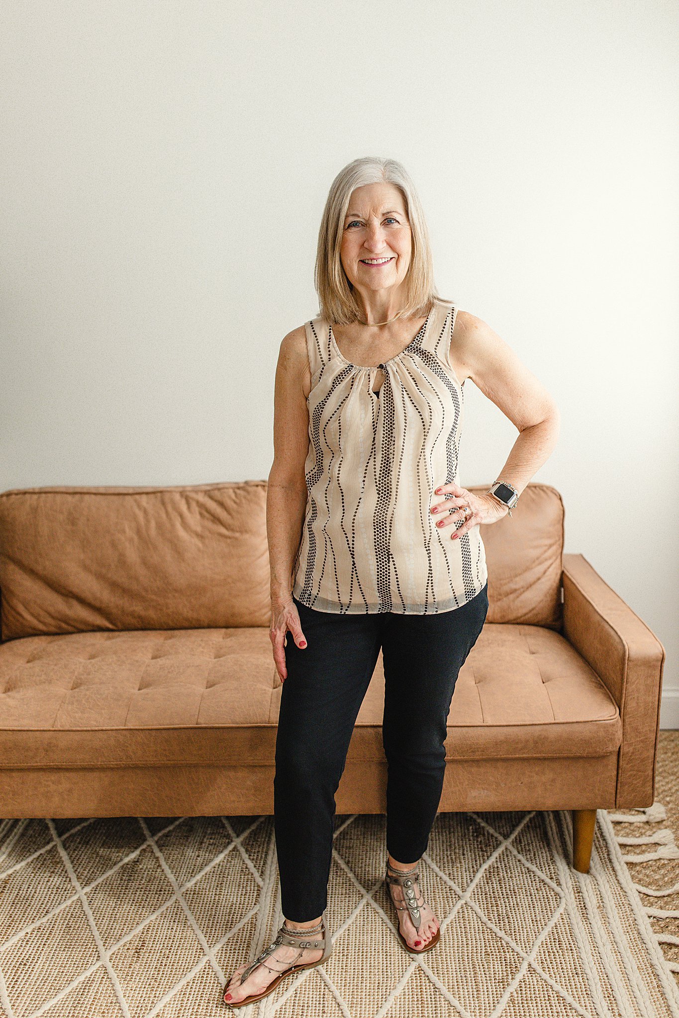 An elderly woman with shoulder-length gray hair, smiling, standing in front of a tan couch in a room with a simple white wall. She's wearing a sleeveless beige top with black dotted lines, black pants with brand photographers in Naperville, IL.