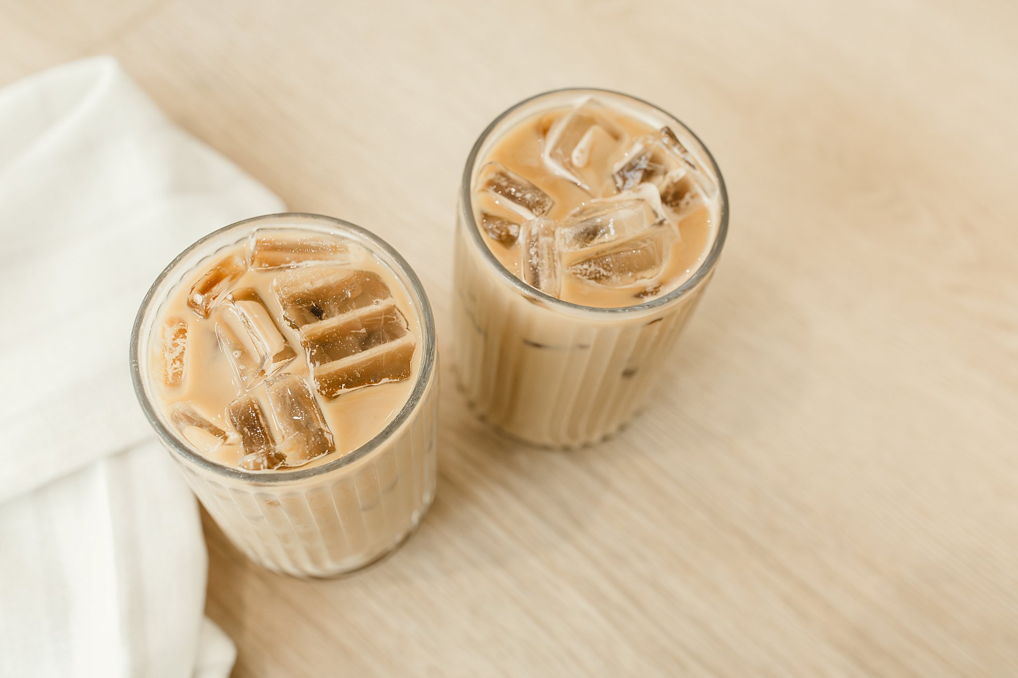 Two glasses of iced coffee on a wooden surface with a white napkin or cloth partially visible on the side for food and drink photographers near me.