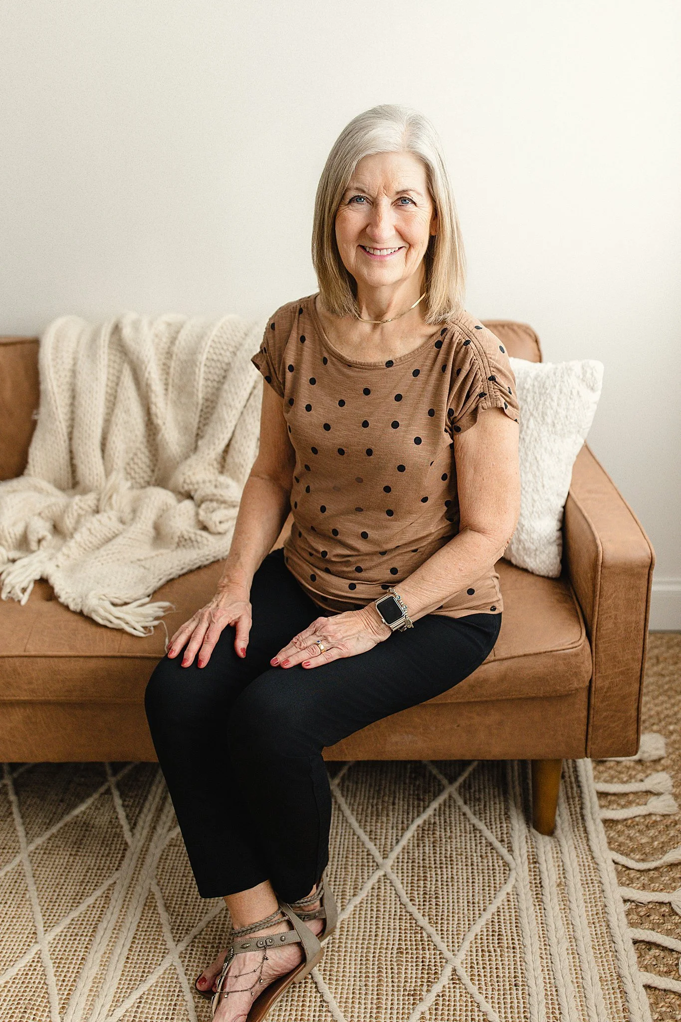 An elderly woman with shoulder-length gray hair, smiling, sitting on a tan sofa, wearing a brown polka dot shirt, black pants, beige sandals, and a smartwatch for headshot photographers near me.