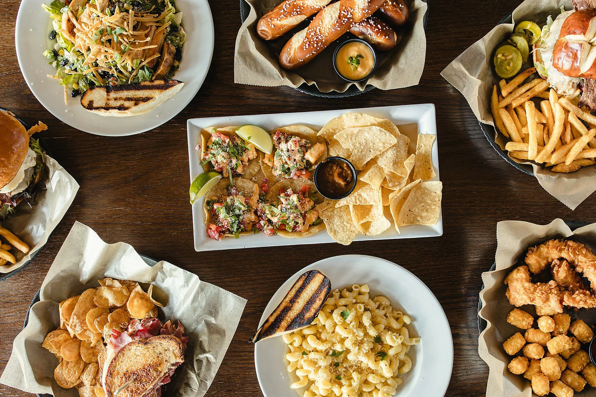 Overhead view of various dishes including a salad, breadsticks with dipping sauce, fries, a burger with toppings, nachos with lime wedges, macaroni and cheese, and fried chicken bites during a food branding session with Ally and B Photography.