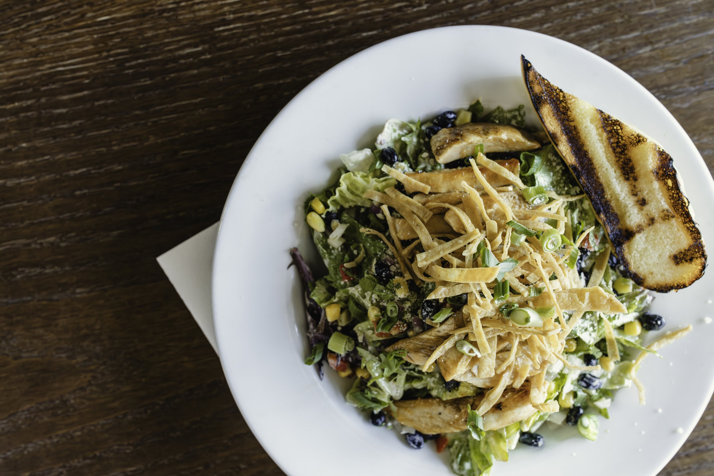 Salad with grilled chicken, shredded cheese, sliced green onions, black beans, corn, and a grilled slice of bread on a white plate for food photography in Chicago, IL.