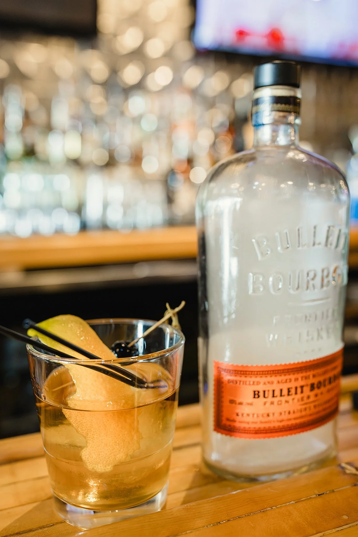 A glass of cocktail garnished with lemon peel and black cherries, with a bottle of Bulleit Bourbon behind it on a wooden bar counter during a bar branding photography session for drink photography of menu items.