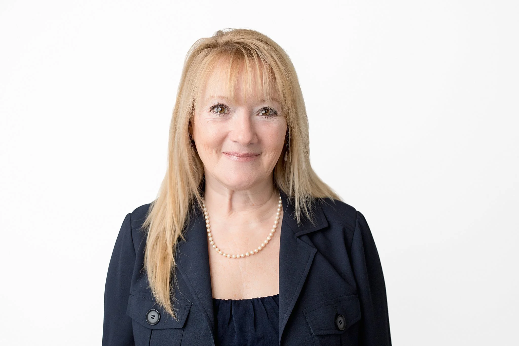 Portrait of a middle-aged woman with blonde hair, wearing a black blazer, black blouse, and a pearl necklace, standing against a plain white background during Naperville headshots.