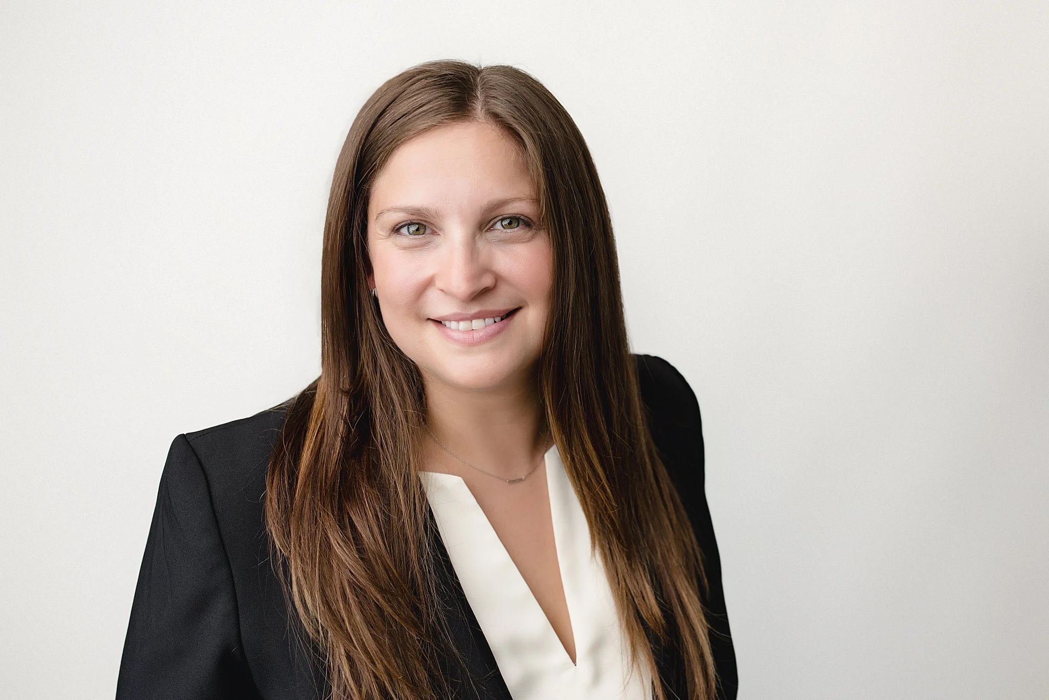Portrait of a woman with long brown hair, wearing a black blazer and white blouse, smiling against a plain white background for Naperville headshot photographer.