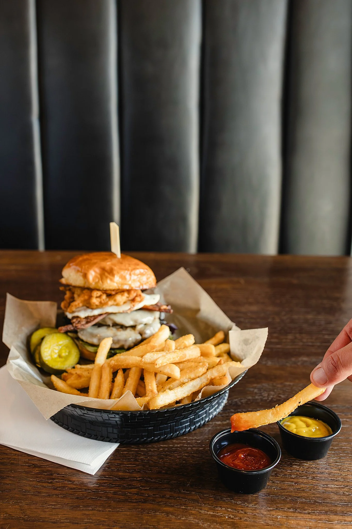 A burger with fried chicken, bacon, cheese, and pickles in a basket with French fries, served with ketchup and mustard on the side, on a wooden table with commercial food photographers near me.