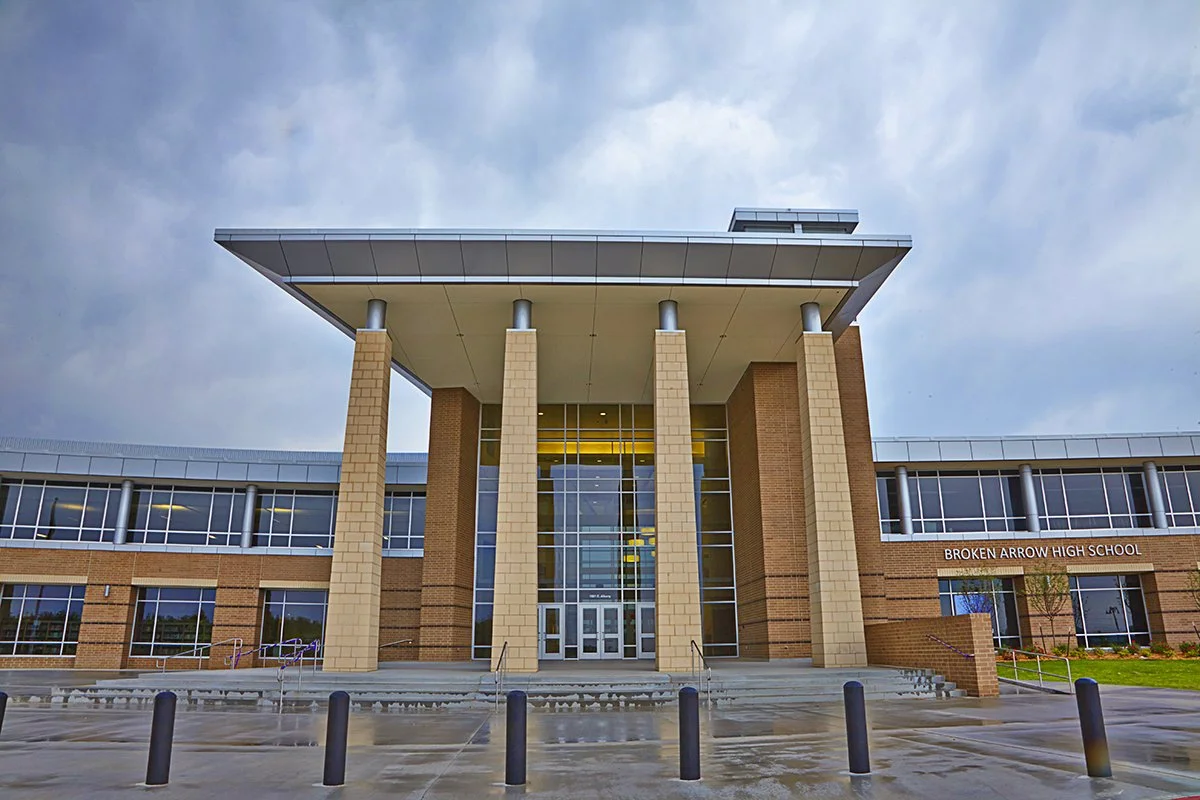 Exterior view of Broken Arrow High School with steps leading to the entrance, large glass door, brick and glass facade, and a cloudy sky.
