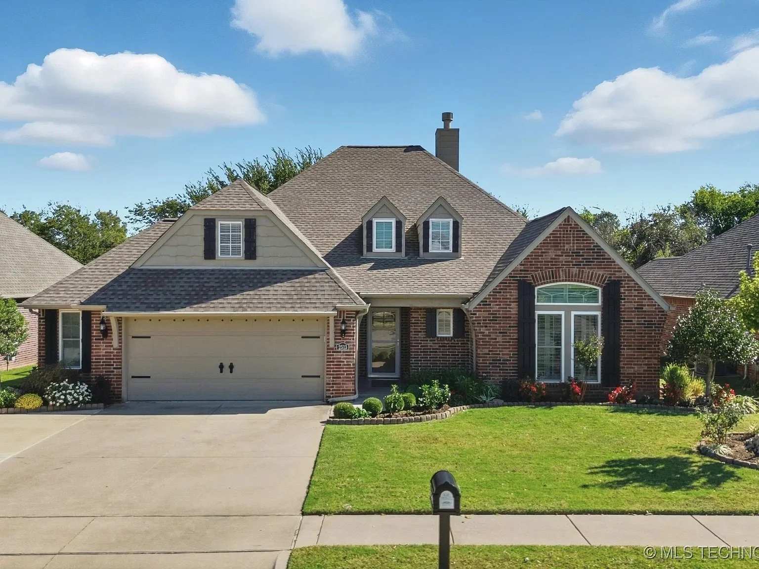 A suburban house with a brick facade, beige siding, and a two-car garage. The yard is well-maintained with green grass, small bushes, and flowers. The sky is blue with a few clouds.