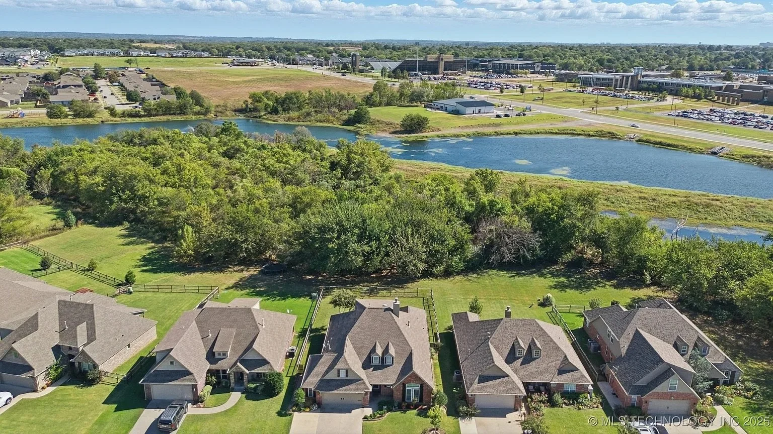 Aerial view of a suburban neighborhood with houses and backyards, a wooded area, and a small pond or lake, with commercial buildings and parking lots in the background.