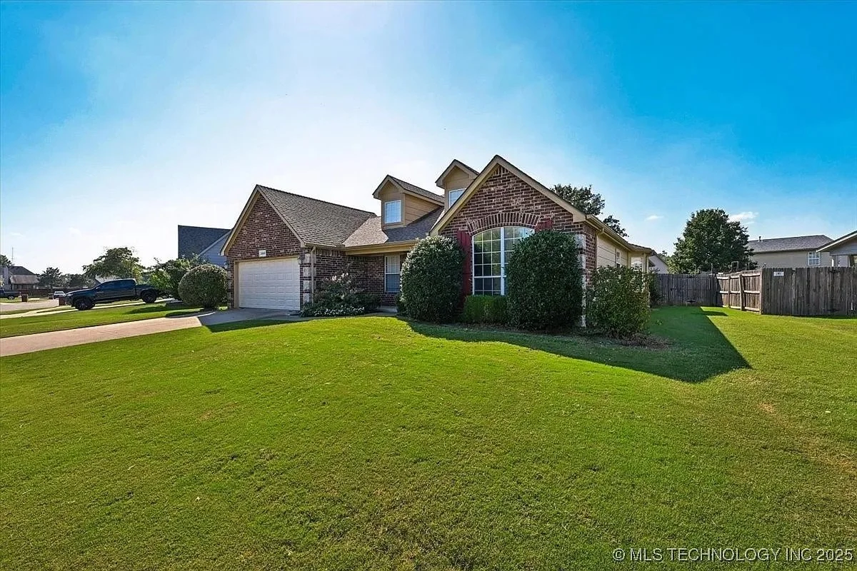 A two-story house with a brick exterior, large front window, and attached garage, surrounded by a well-maintained green lawn and bushes, under a clear blue sky.