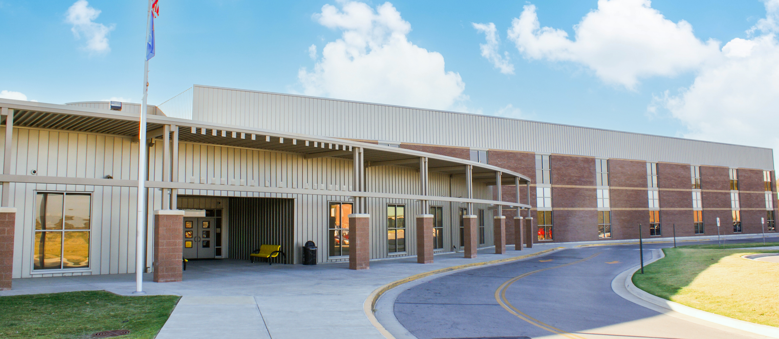 Exterior of a modern school building with a curved driveway, flagpole, bench, and green lawn, under a partly cloudy sky.