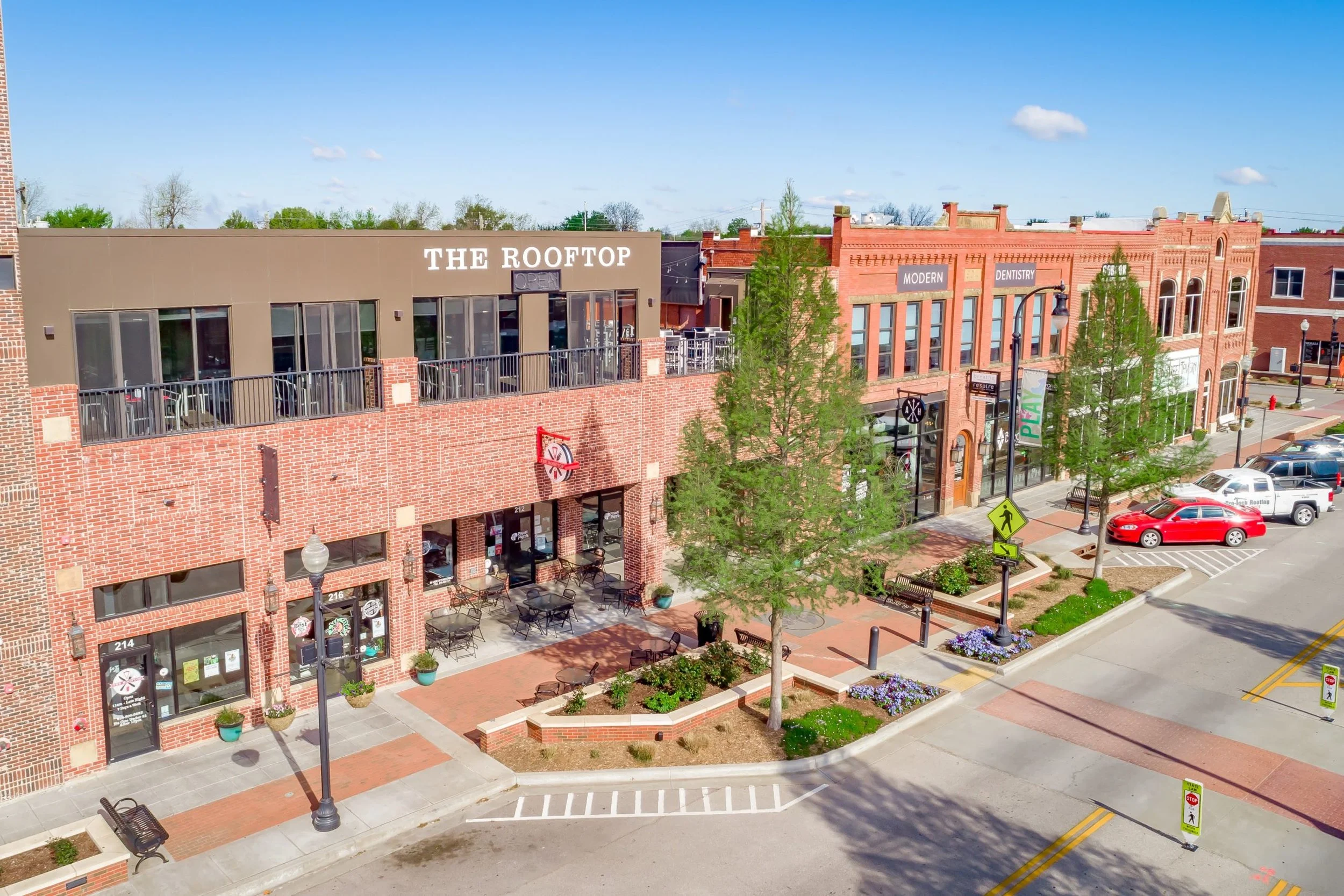 A street view of a shopping area with brick buildings, storefronts, trees, parked cars, benches, and pedestrian crosswalk signs under a blue sky with a few clouds.