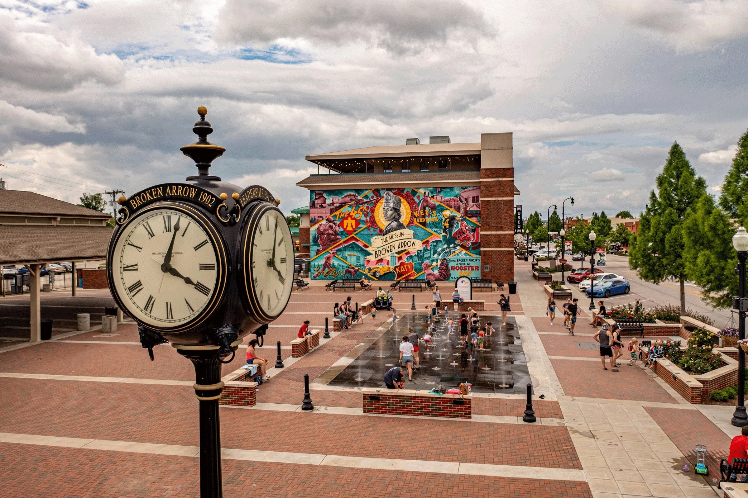 Public plaza with a large clock in the foreground, a colorful mural on a building in the background, children playing in a water fountain, and people walking and sitting on benches, with trees and parked cars along the street.