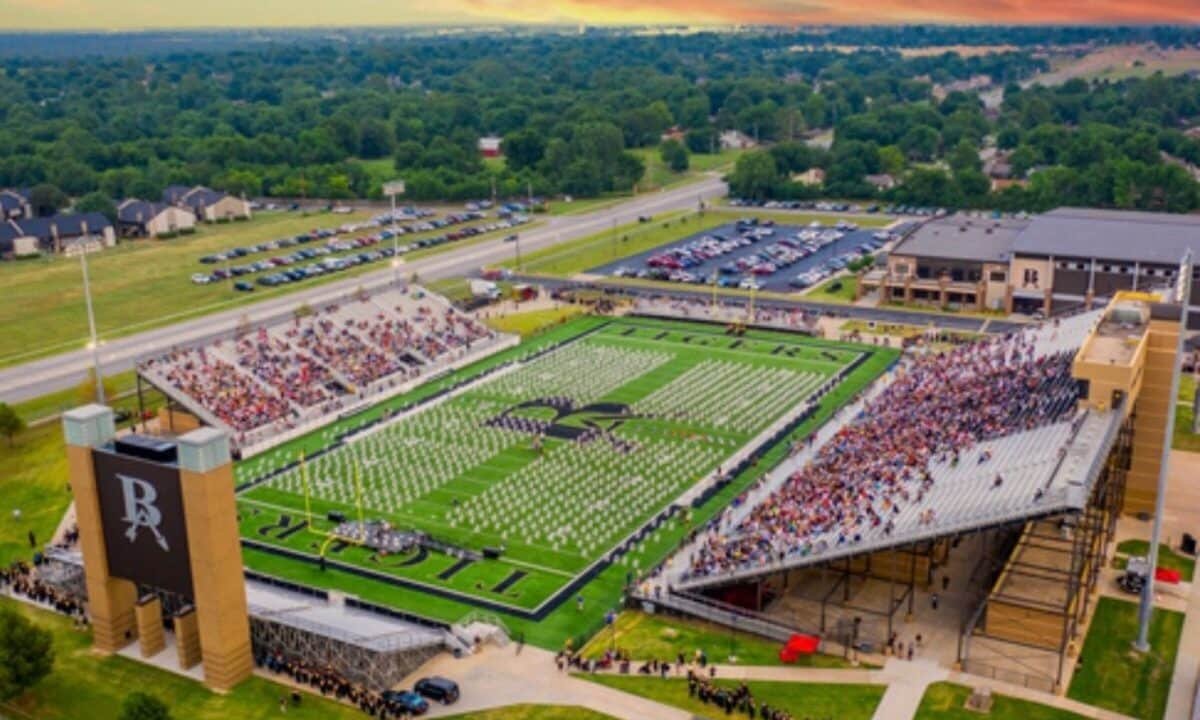 Aerial view of a university football stadium with a crowd of spectators, green turf with a large logo in the center, and adjacent parking lots and residential area.