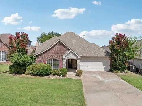 Front view of a brick house with a gray roof, two car garage, and landscaped yard with bushes and trees.
