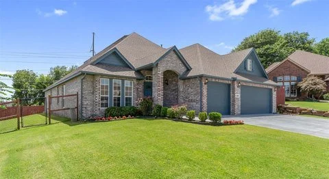 A brick suburban house with a three-car garage, front lawn, and a fence on the left, under a blue sky.