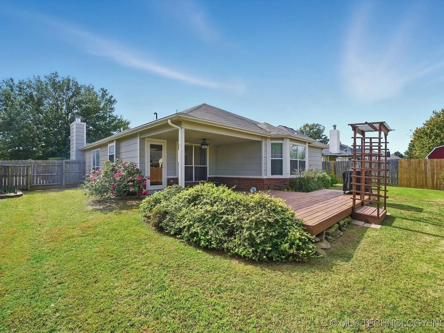 Backyard of a house with a wooden deck, shrubbery, a flowering bush, a grill, and a pergola, enclosed by a wooden fence under a clear blue sky.
