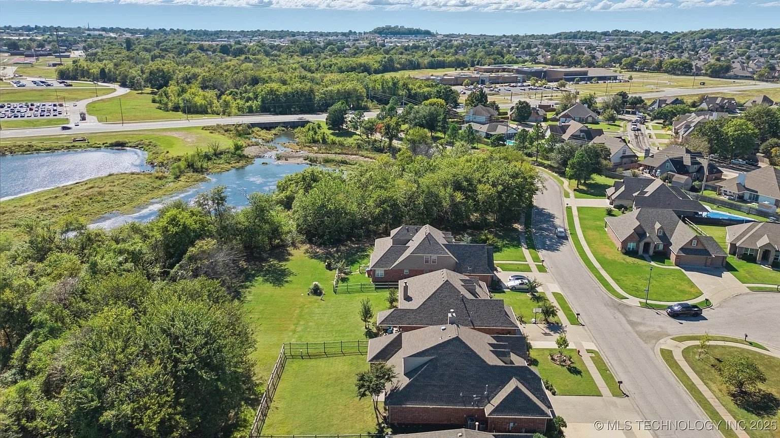 Aerial view of a suburban neighborhood near a pond, with houses, green lawns, trees, and a parking lot in the background.