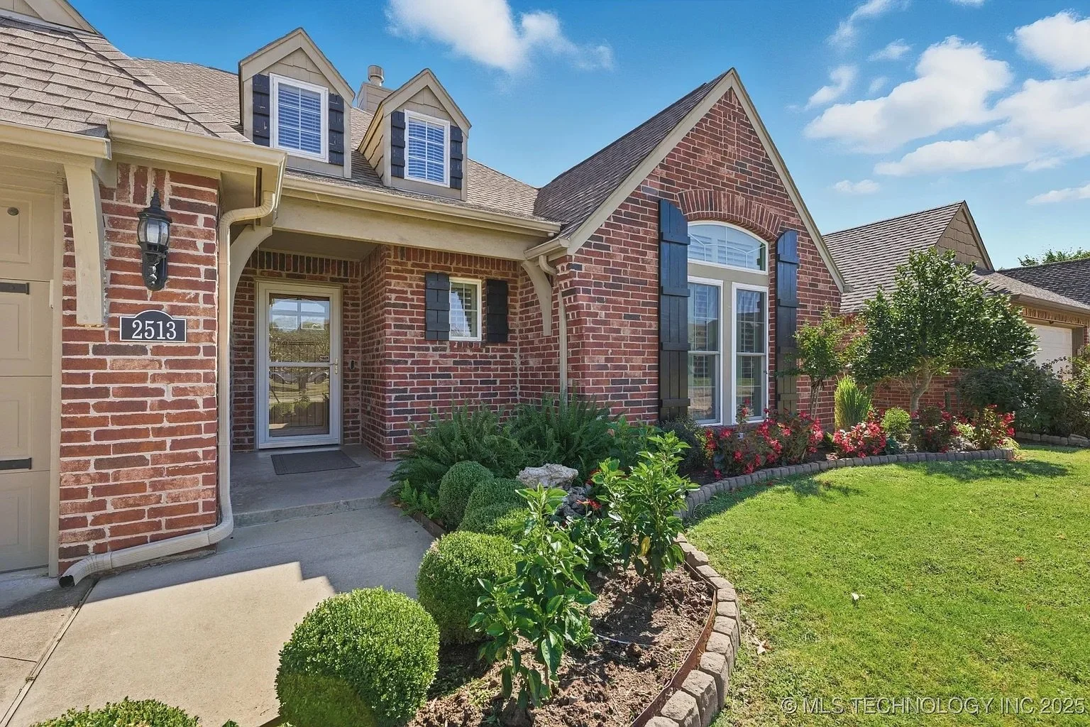Front of a brick house with a garden, trees, and a lawn, under a blue sky.