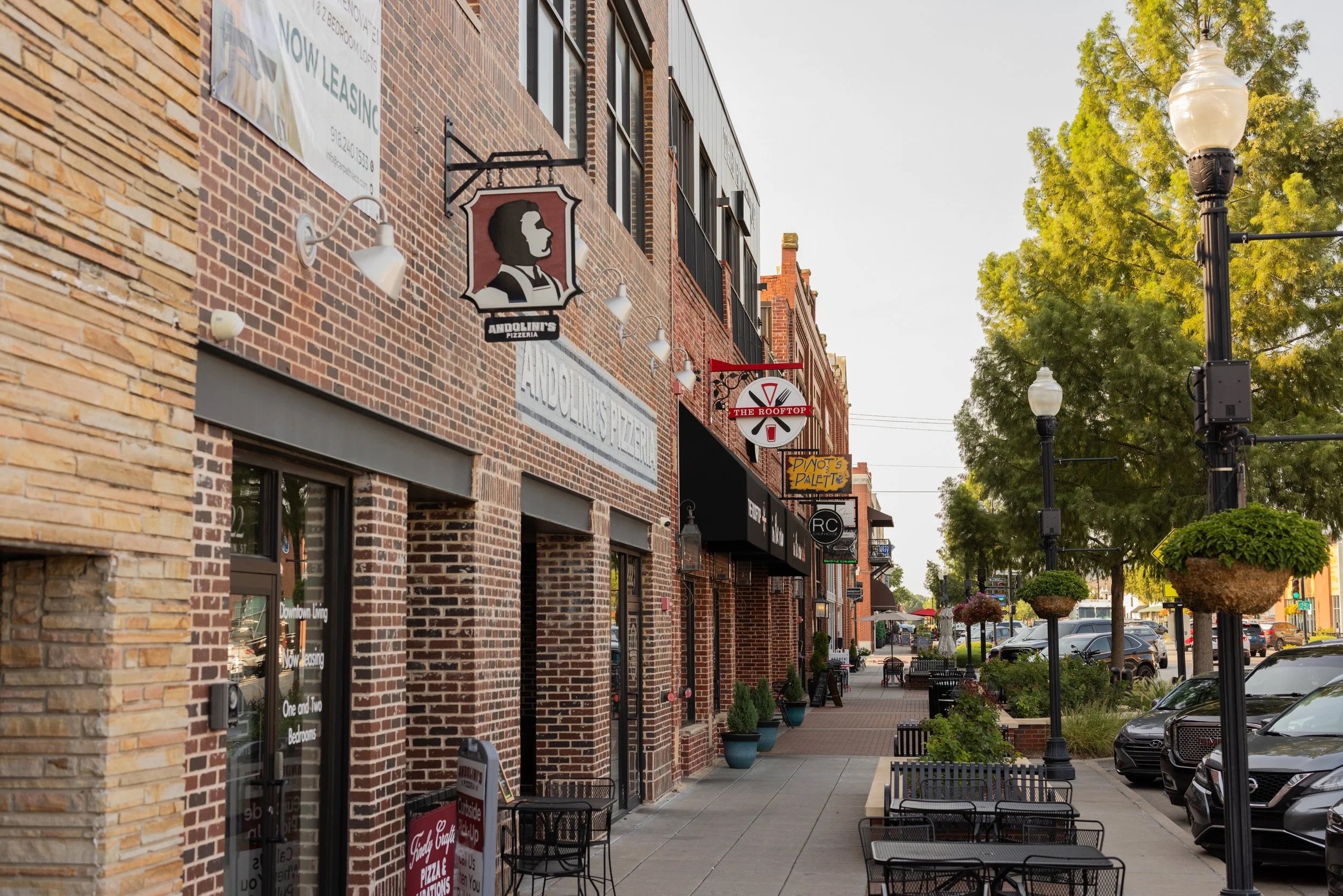 A sidewalk scene in an urban shopping area with brick storefronts and outdoor seating, along with street lamps, trees, and parked cars.